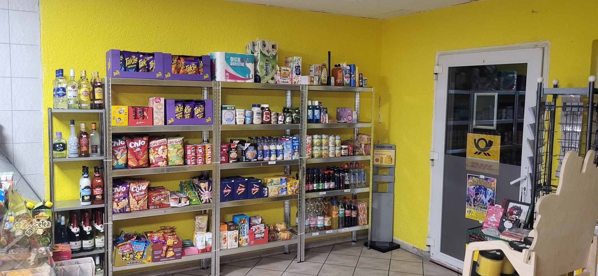 Ein kleiner Laden mit Regalen vor einer leuchtend gelben Wand, gefüllt mit Snacks, Konserven und Getränken. Eine Glastür auf der rechten Seite führt in einen weiteren Bereich