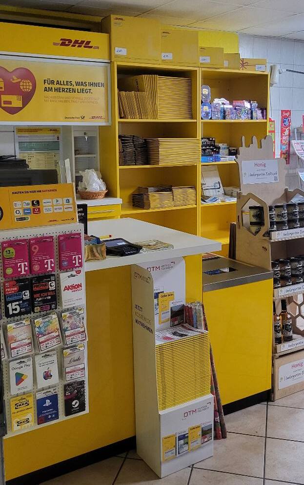 Das Innere einer DHL-Postfiliale mit gelben Regalen, die mit Briefumschlägen und Paketen gefüllt sind. Auf einem Schalter liegen verschiedene Karten und ein Schild aus